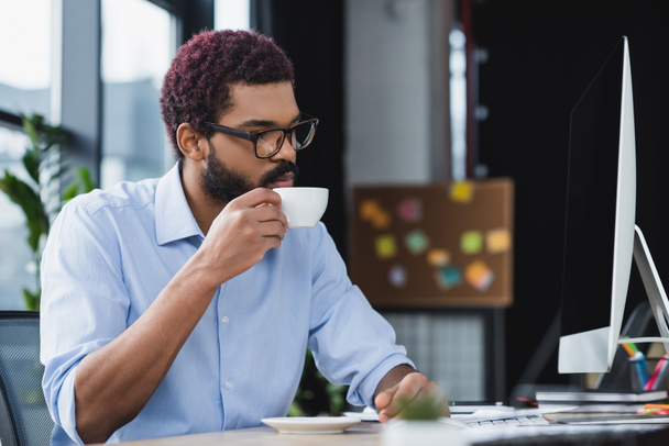 若いアフリカ系アメリカ人ビジネスマンがオフィスのコンピュータモニターの近くでカップを保持  - 写真・画像
