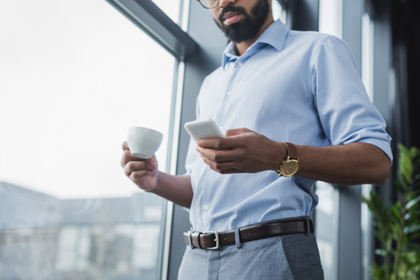 Alacsony látószögű kilátás afro-amerikai üzletember használja a mobiltelefon és a kezében kávéscsésze az irodában  - Fotó, kép