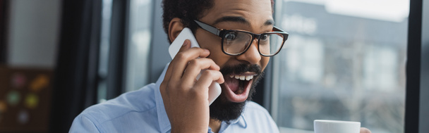 Étonné homme d'affaires afro-américain parlant sur téléphone portable et tenant la tasse dans le bureau, bannière  - Photo, image