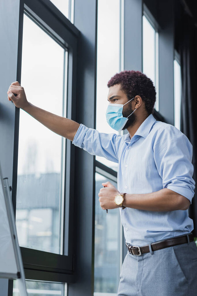 Giovane uomo d'affari afroamericano in maschera medica in piedi vicino alla finestra e lavagna a fogli mobili  - Foto, immagini