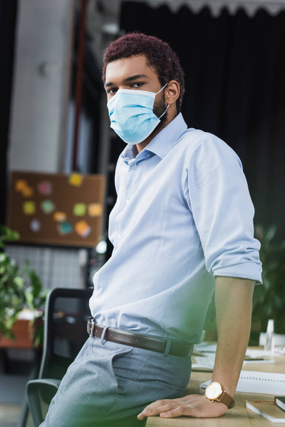 Junger afrikanisch-amerikanischer Geschäftsmann mit medizinischer Maske blickt im Büro in die Kamera  - Foto, Bild