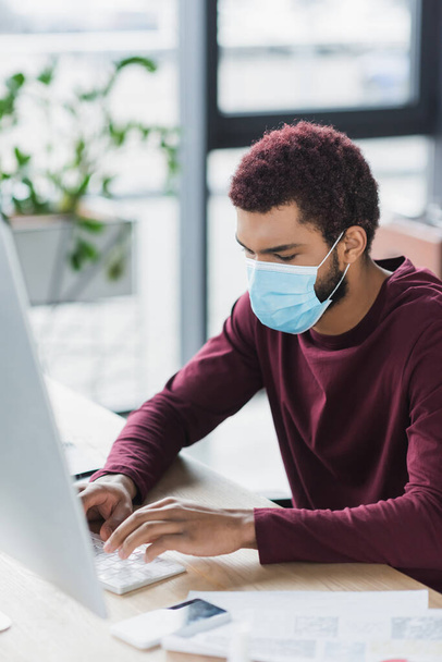 African american businessman in medical mask using computer keyboard near blurred monitor in office - Photo, Image