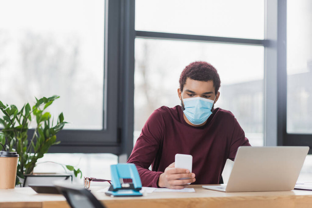 African american businessman in protective mask using mobile phone near laptop and coffee to go in office  - Photo, Image
