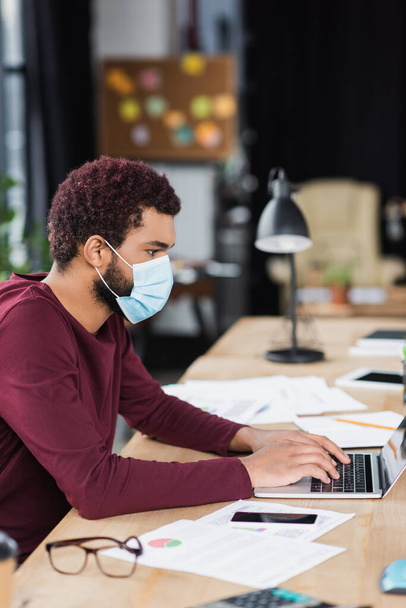 Side view fiatal afro-amerikai üzletember orvosi maszk segítségével laptop közelében papírok és szemüveg az irodában  - Fotó, kép