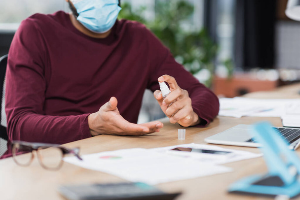 Ausgeschnittene Ansicht eines afrikanisch-amerikanischen Geschäftsmannes in medizinischer Maske mit Händedesinfektionsmittel in der Nähe von Geräten im Büro  - Foto, Bild