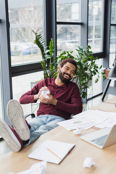 Glimlachende Afro-Amerikaanse zakenman houdt verfrommeld papier in de buurt van laptop in het kantoor  - Foto, afbeelding