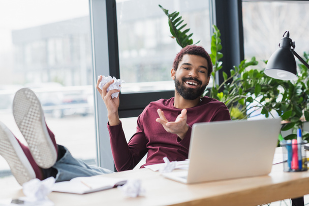 Empresario afroamericano positivo sosteniendo papel arrugado durante videollamada en la computadora portátil en la oficina  - Foto, Imagen