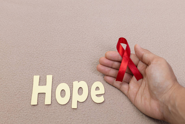 A hand holding a red awareness ribbon and the word hope on a brown background - Φωτογραφία, εικόνα