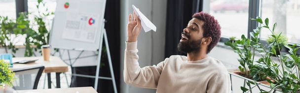 Vidám afro-amerikai üzletember kezében papír repülőgép az irodában, banner  - Fotó, kép