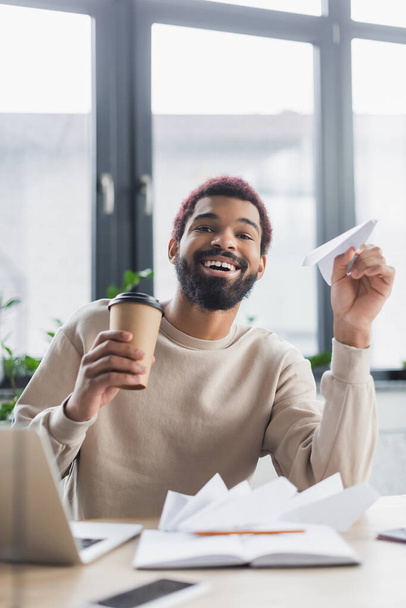 Pozitivní africký americký podnikatel drží kávu jít a papírové letadlo v blízkosti notebooku v kanceláři  - Fotografie, Obrázek