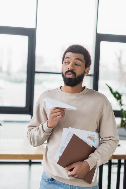Joven empresario afroamericano sosteniendo avión de papel y documentos en la oficina  - Foto, imagen