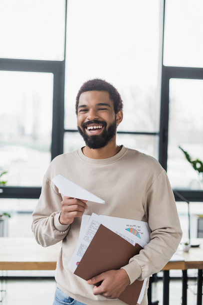 Χαρούμενος Αφροαμερικάνος επιχειρηματίας με χάρτινο αεροπλάνο και έγγραφα στο γραφείο  - Φωτογραφία, εικόνα