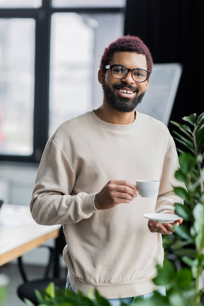 Happy young african american businessman in eyeglasses holding cup of coffee in office  - Foto, imagen