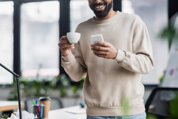 Cropped view of smartphone and cup in hands of blurred african american businessman in office  - Foto, Imagen