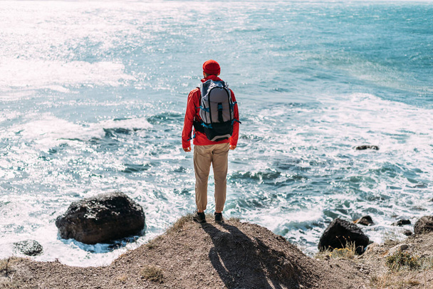 A man with a backpack on the background of the sea, rear view. A man in tourist gear on the background of the sea. The concept of travel and an active lifestyle. A traveler on the seashore. Copy space - Фото, зображення