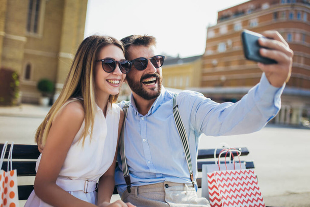 Retrato de hermosa pareja joven sonriente sentada en un banco después de ir de compras hacer foto selfie. - Foto, Imagen