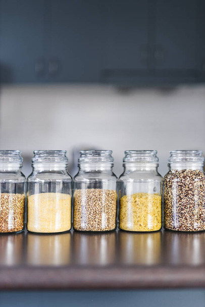 clear pantry jars with different types of grains and legumes including quinoa  lentils buckwheat and barley on benchtop with kitchen bokeh, simple ingredients concept - Фото, зображення