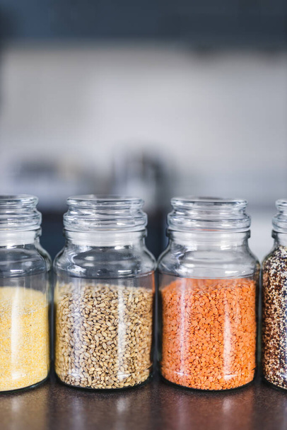 clear pantry jars with different types of grains and legumes including quinoa  lentils buckwheat and barley on benchtop with kitchen bokeh, simple ingredients concept - Foto, Imagen