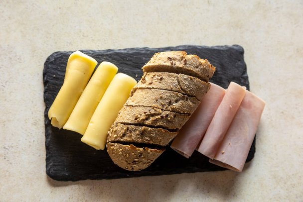A top view of a black board with sliced bread, sliced ham, and cheese - Фото, изображение