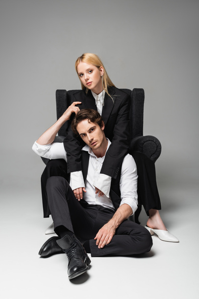 blonde woman in armchair embracing elegant man sitting on floor on grey - 写真・画像