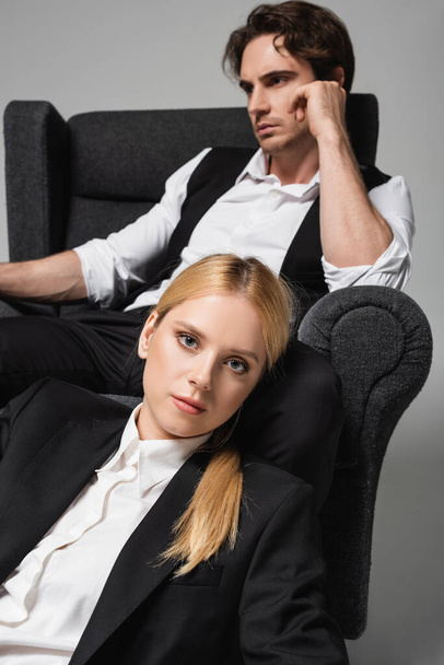 elegant blonde woman looking at camera near blurred man sitting in armchair isolated on grey - Fotografie, Obrázek
