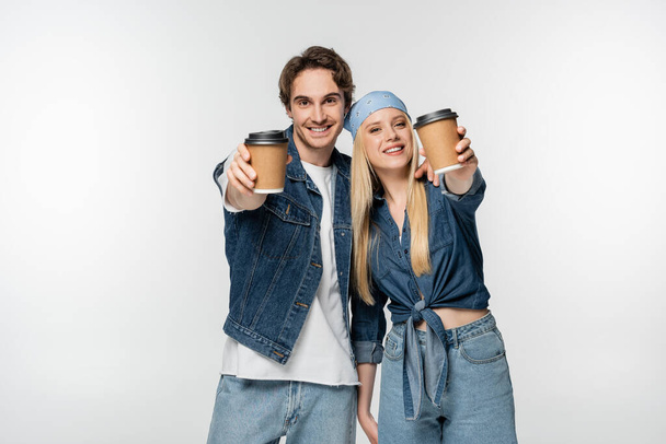 smiling couple in trendy denim clothes showing paper cups isolated on white - Photo, Image