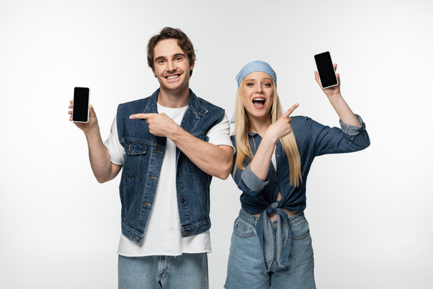 happy stylish couple pointing at mobile phones with blank screen isolated on white - 写真・画像