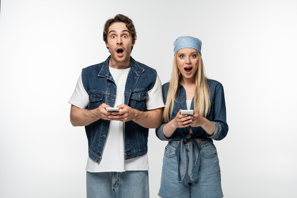 astonished couple in trendy denim clothes looking at camera while using cellphones isolated on white - Fotografie, Obrázek