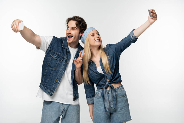 excited man showing victory gesture while taking selfie on smartphones together with woman isolated on white - Valokuva, kuva