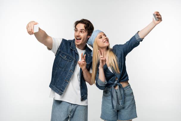 happy couple in denim clothing showing victory sings while taking selfie isolated on white - Foto, Imagem