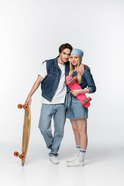trendy couple with longboard and penny board embracing and looking at camera on white - Zdjęcie, obraz
