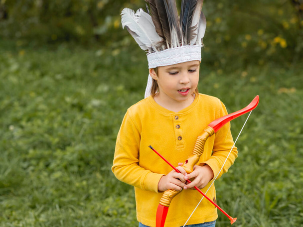 Little boy is playing American Indian on field. Kid has handmade headdress made of feathers and bow with arrows. Costume role play. Outdoor leisure activity. Fall season. - Фото, зображення