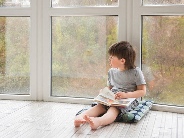 Le gamin s'assoit par terre et lit un livre. Loisirs intellectuels pour les enfants pendant qu'il pleut dehors. - Photo, image