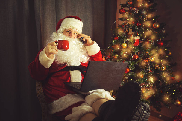 Babbo Natale autentico sta lavorando su un computer portatile e sta parlando al telefono. Interni domestici. In previsione di Natale e Capodanno.  - Foto, immagini