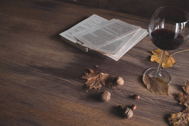 Still life with red wine and book. Nuts, autumn leaves, glass of wine and open book on wooden table. Lifestyle concept. Cozy home interior. Hazelnut, walnut and wine. Vintage still life. - Photo, Image