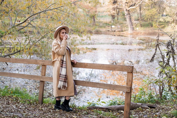 Güz parkında, bej ceketli ve şapkalı çekici bir kadın gölün kenarında kahve içiyor. - Fotoğraf, Görsel