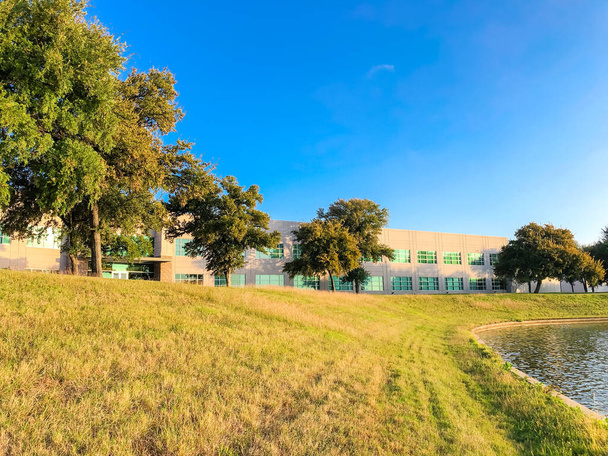 Low angle view from a steep slope hill lawn of lakeside business park in Lewisville, Texas, America. Waterfront corporate buildings with matured trees under blue cloud sky, early morning - Photo, Image