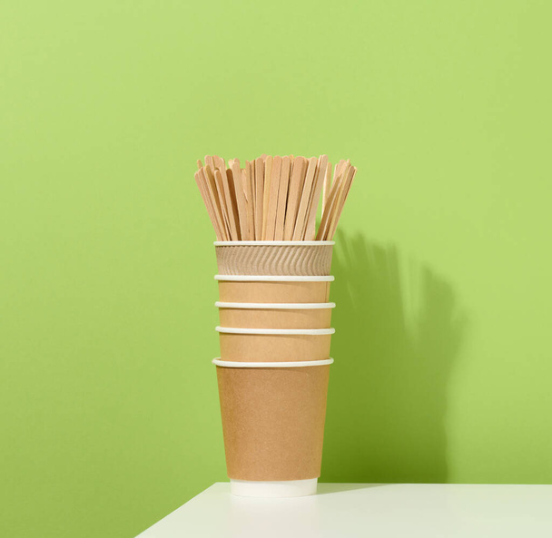 vasos de cartón de papel marrón y palos de madera revolviendo sobre una mesa blanca, fondo verde. Vajilla ecológica - Foto, imagen