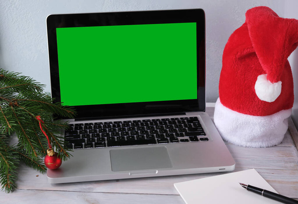 Christmas sales and online shopping for gifts.Laptop with green blank screen, Santa hat, fir branches and red ball on wooden background. Selection presents of for new year.List of purchases, gifts.Selective focus - Foto, Imagem