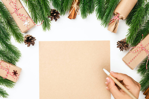 Flat lay, mock-up wish list and female hand writing checklist on table with decorations of fir branches and gift boxes - 写真・画像