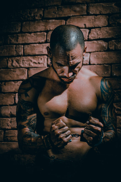 Ένας μελαχρινός άντρας με κοντό κούρεμα και τατουάζ κάθεται στη φυλακή με τα χέρια του με χειροπέδες. Τιμωρία για φόνο.. - Φωτογραφία, εικόνα