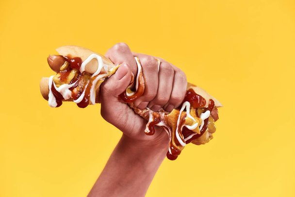 Female hand squeezing out junk food, hot dog with mustard and ketchup isolated over yellow background - Zdjęcie, obraz