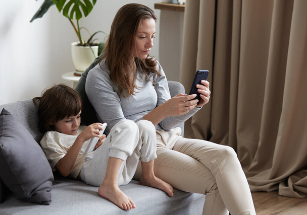 Focused modern mother and son kid chatting surfing internet use smartphone sitting on couch together - Photo, Image