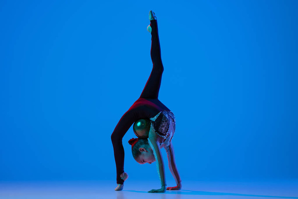 Ganzkörperporträt eines kleinen Mädchens, professionelles rhythmisches Turner-Training, das isoliert vor blauem Hintergrund in Neon auftritt - Foto, Bild