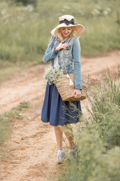 a young blonde woman with blue eyes in a straw hat and denim dress is holding a straw bag with flowers in the Park on a summer day. The concept of romance - Φωτογραφία, εικόνα