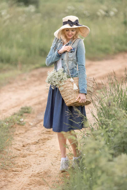a young blonde woman with blue eyes in a straw hat and denim dress is holding a straw bag with flowers in the Park on a summer day. The concept of romance - Photo, Image