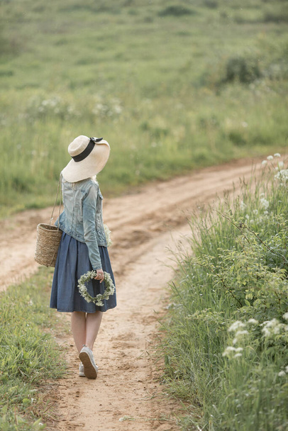 Rear view young woman in denim dress straw hat and bag holding a wreath of forest flowers standing in the way of the forest - Φωτογραφία, εικόνα