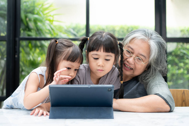 Ασιατική γιαγιά με τα δύο εγγόνια της να διασκεδάζουν και να παίζουν online εκπαιδευτικά παιχνίδια με ένα ψηφιακό tablet στο σπίτι στο σαλόνι. Έννοια της online εκπαίδευσης και φροντίδας από τους γονείς. - Φωτογραφία, εικόνα