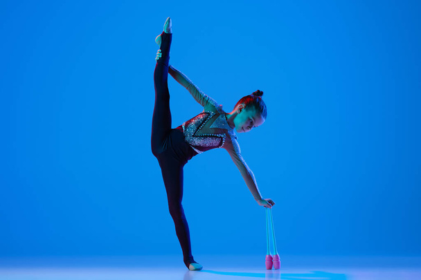 Portrait of little girl, professional rhythmic gymnast performing with gymnastic clubs isolated over blue background in neon - Foto, Imagem