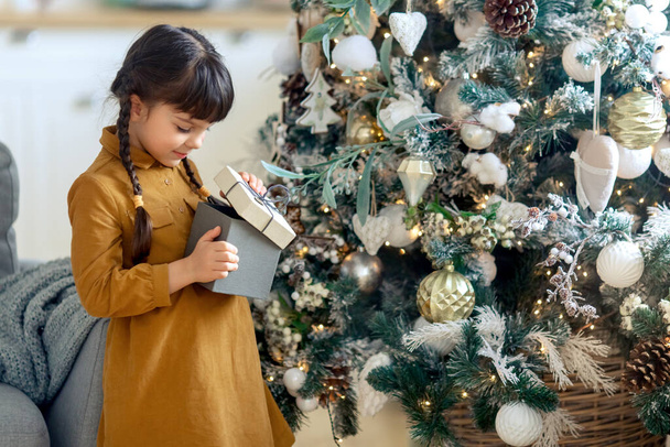 Cute girl 5 years old in a yellow dress with a gift box in her hands standing near the Christmas tree. New year - Foto, Bild